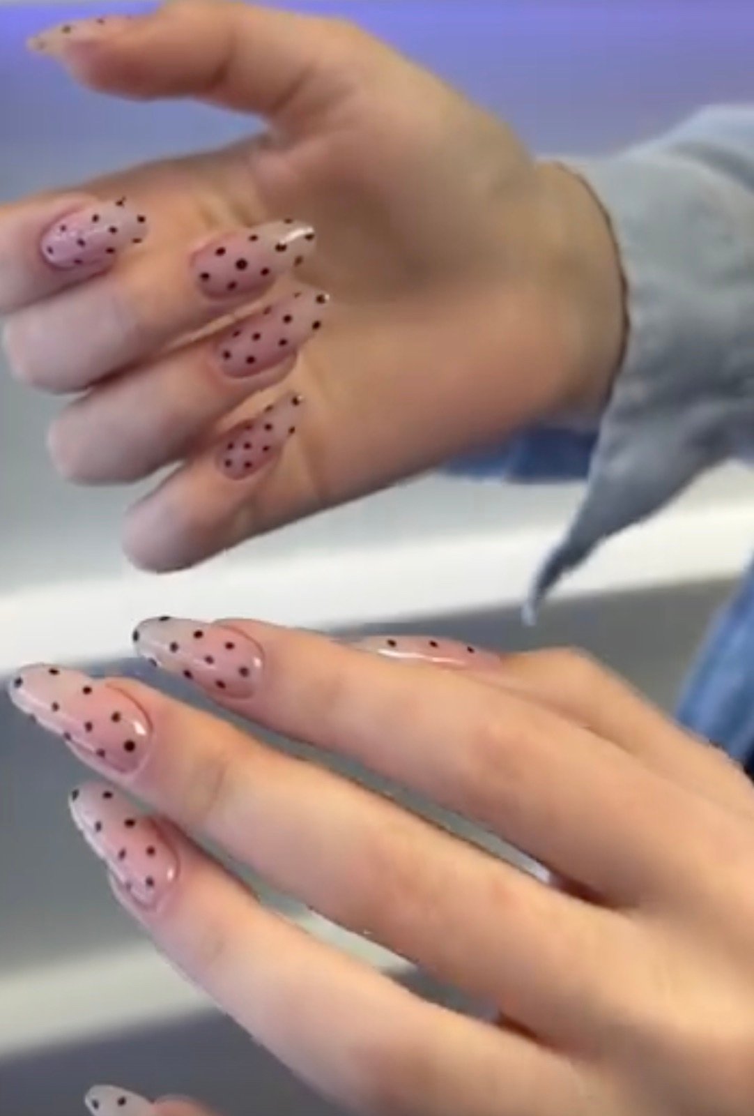 Nail design inspiration for russian manicure: Two hands with long almond-shaped nails featuring a light pink base and black polka dot design, glossy finish