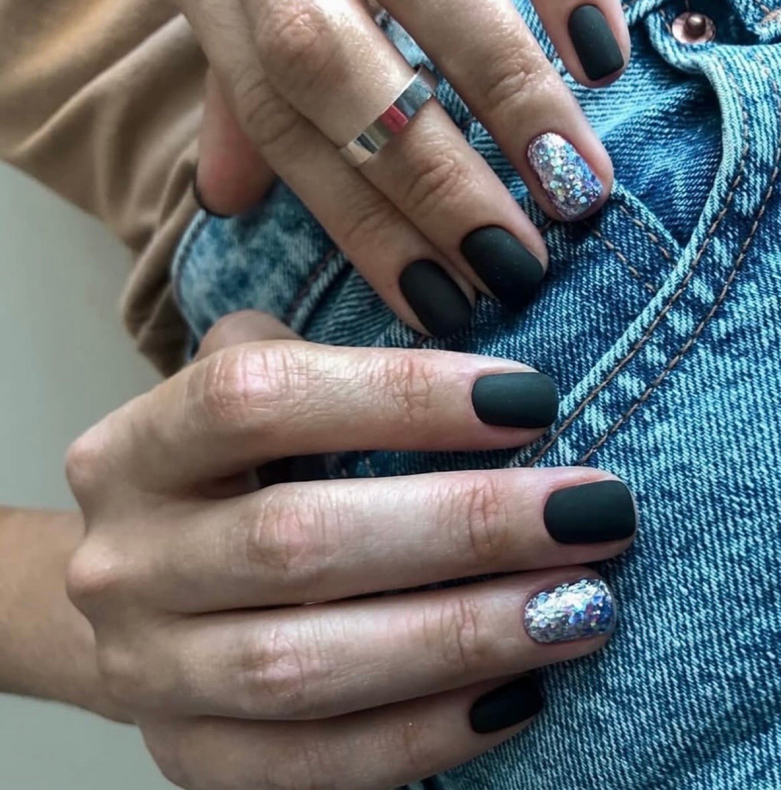 Nail design inspiration for russian nail salon near me: Close-up of hands with matte black nail polish and glitter accents on two fingernails against denim fabric