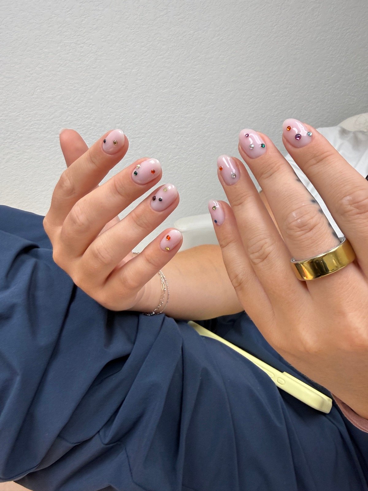 Nail design inspiration for russian nails near me: Close-up of a hand with light pink manicure featuring small gemstone accents on each nail, wearing gold ring and dark blue top